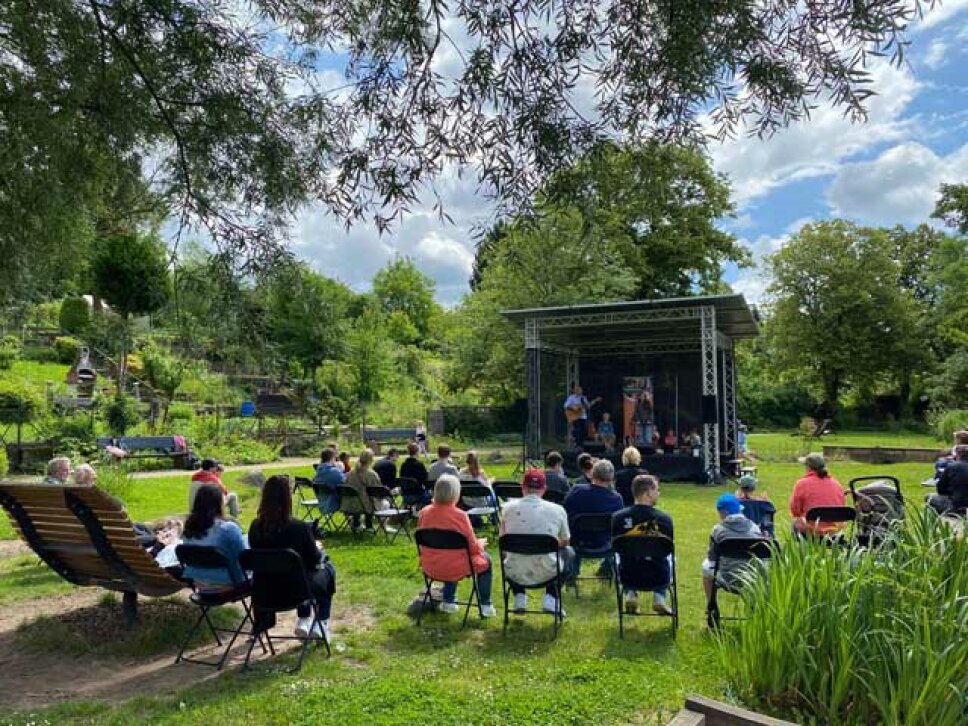 Menschen sitzen in einem Park vor einer Bühne und hören einem Konzert zu